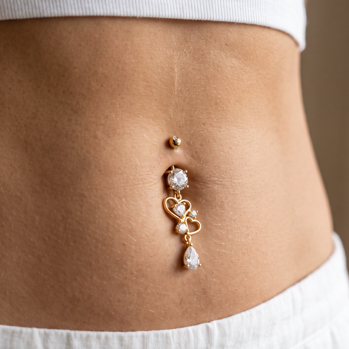 A close-up of a person's midsection wearing a gold belly button ring with clear rhinestone accents, styled with a white ribbed crop top and white pants.