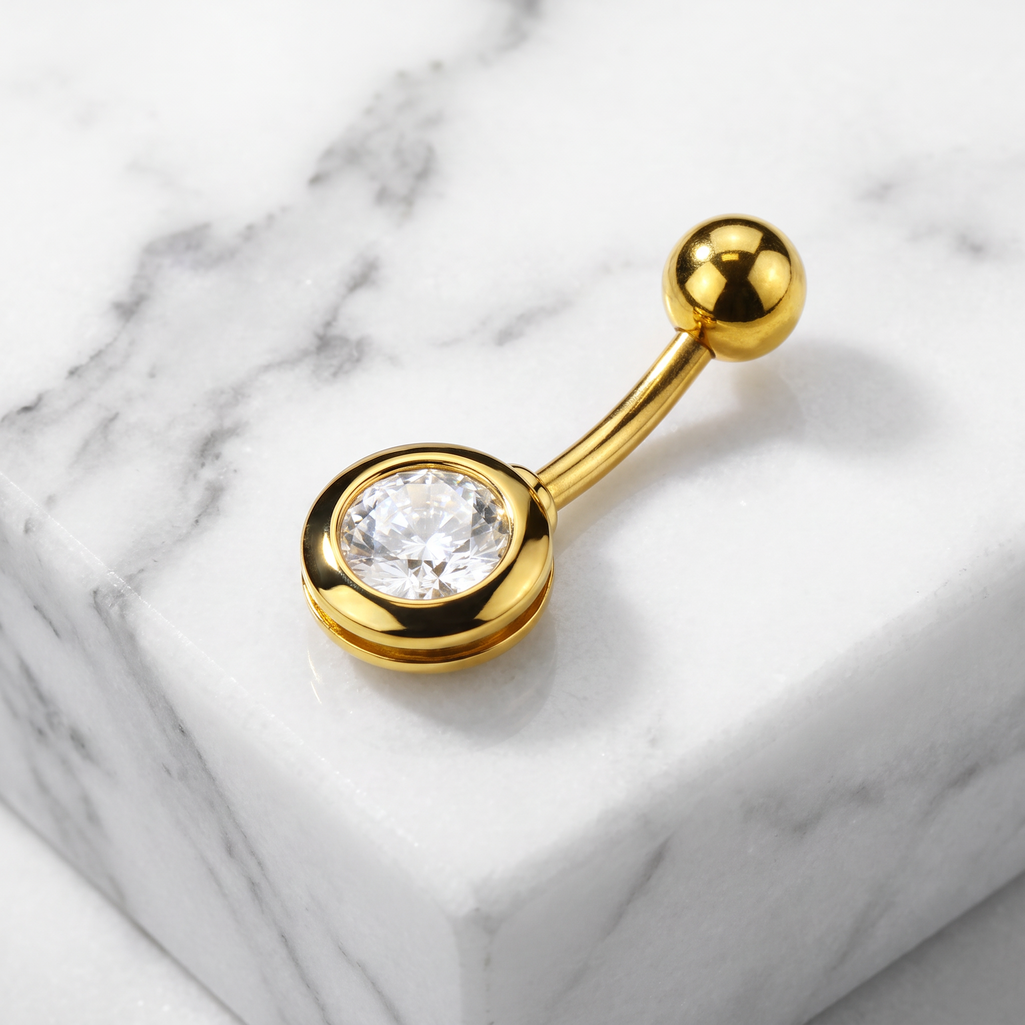 A gold-plated curved barbell navel piercing stud with a round bezel-set clear gemstone, displayed on a white marble block.