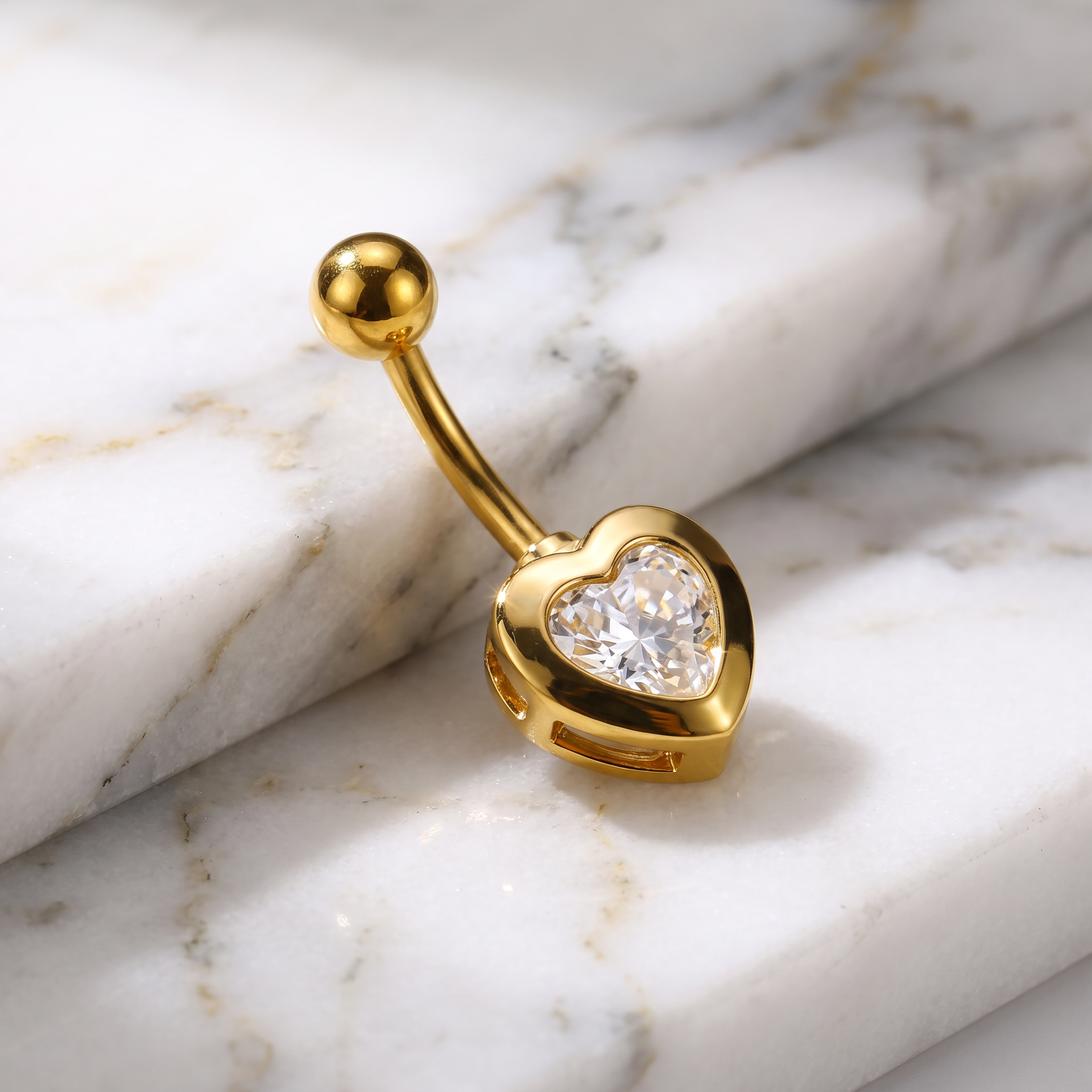 Gold-plated belly button ring with a heart-shaped bezel containing a clear heart-shaped gemstone, displayed on a white marble background.