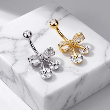 Two bow-shaped belly button rings with clear rhinestone accents, one in silver and one in gold, displayed on a white marble block.