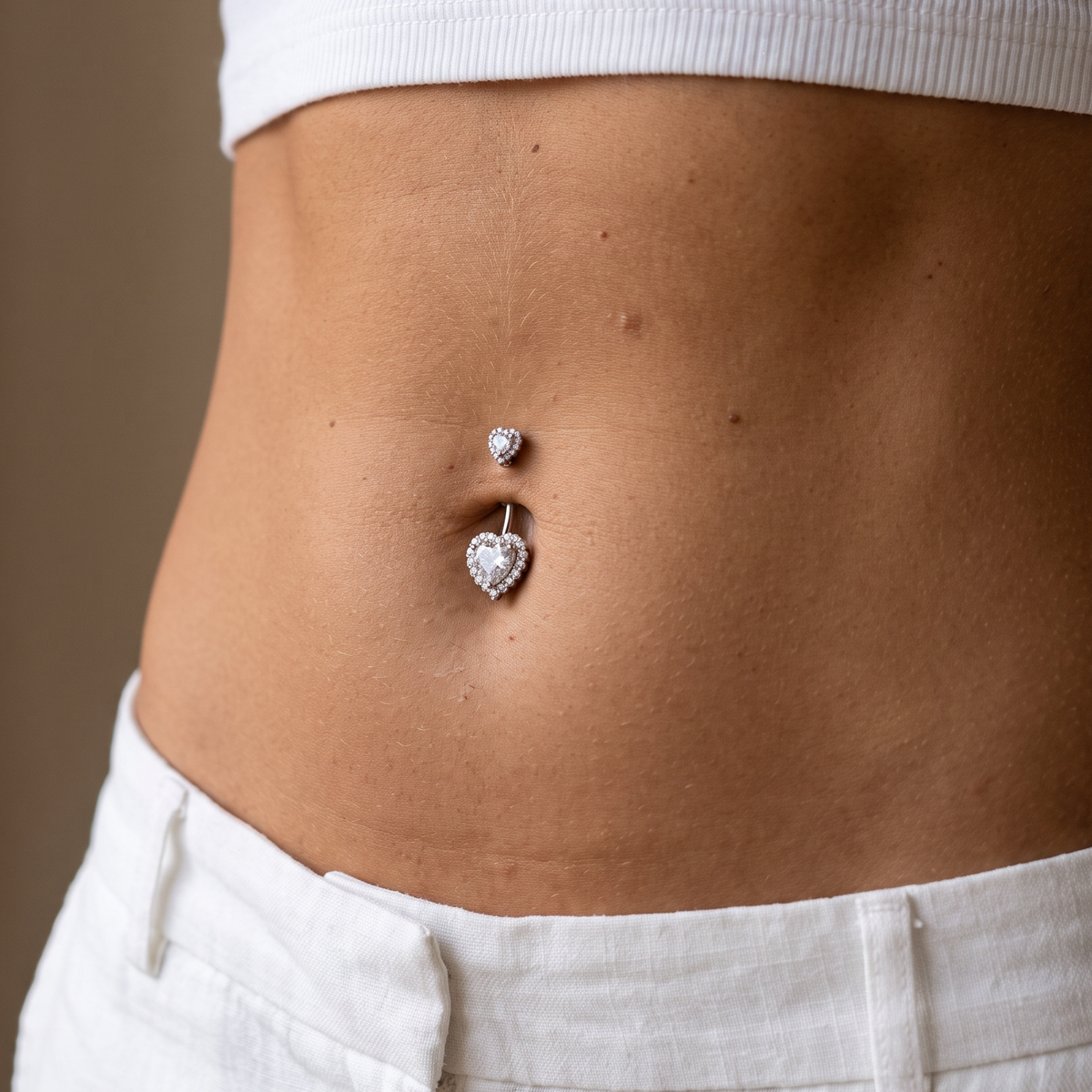 A close-up of a person's midsection wearing a silver belly button ring with a small round stud and a heart-shaped pavé charm, paired with a white ribbed crop top and white pants.