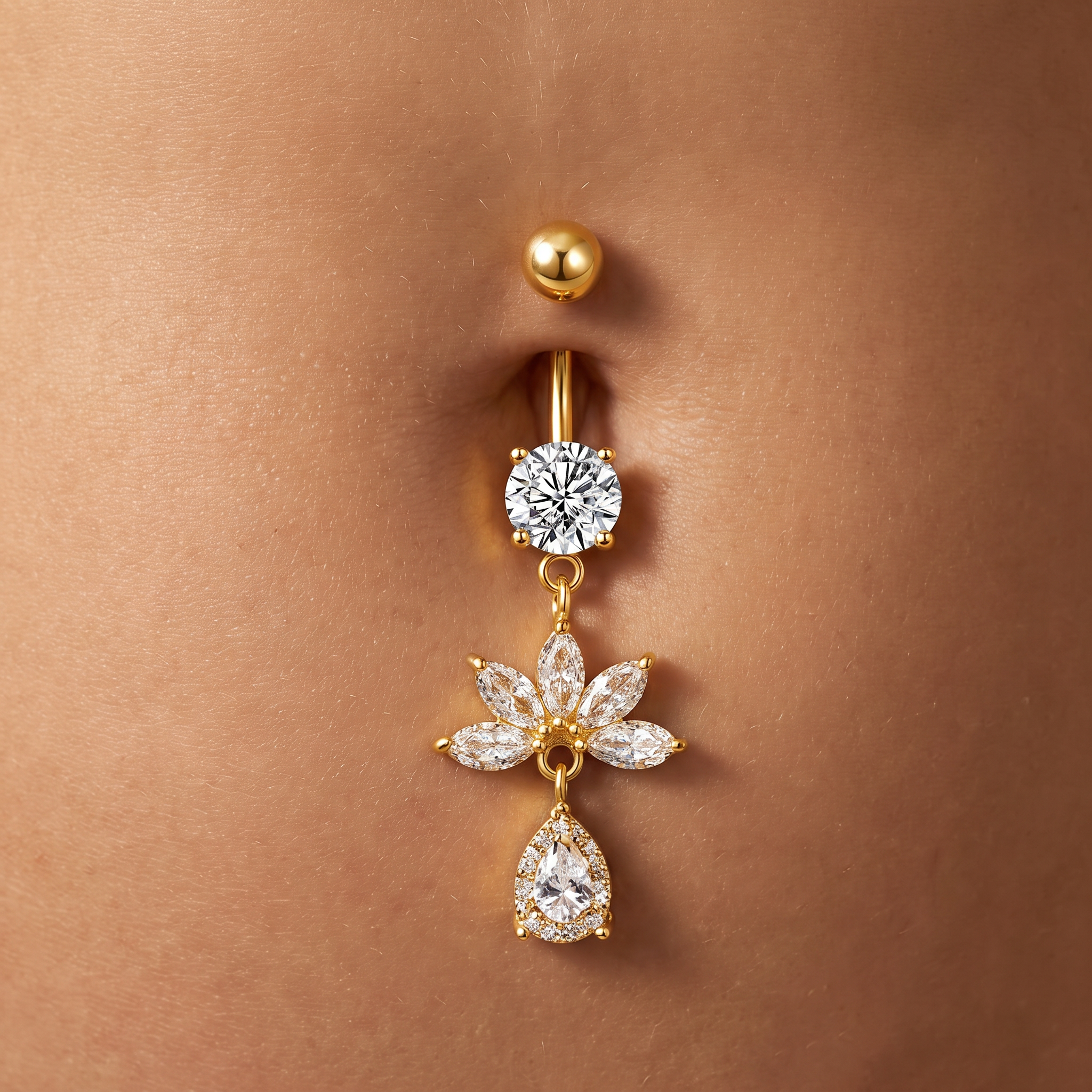 A gold-plated belly button ring with a round bezel-set clear gemstone and a dangling flower charm with pear-cut clear gemstones, shown worn on a person's abdomen.