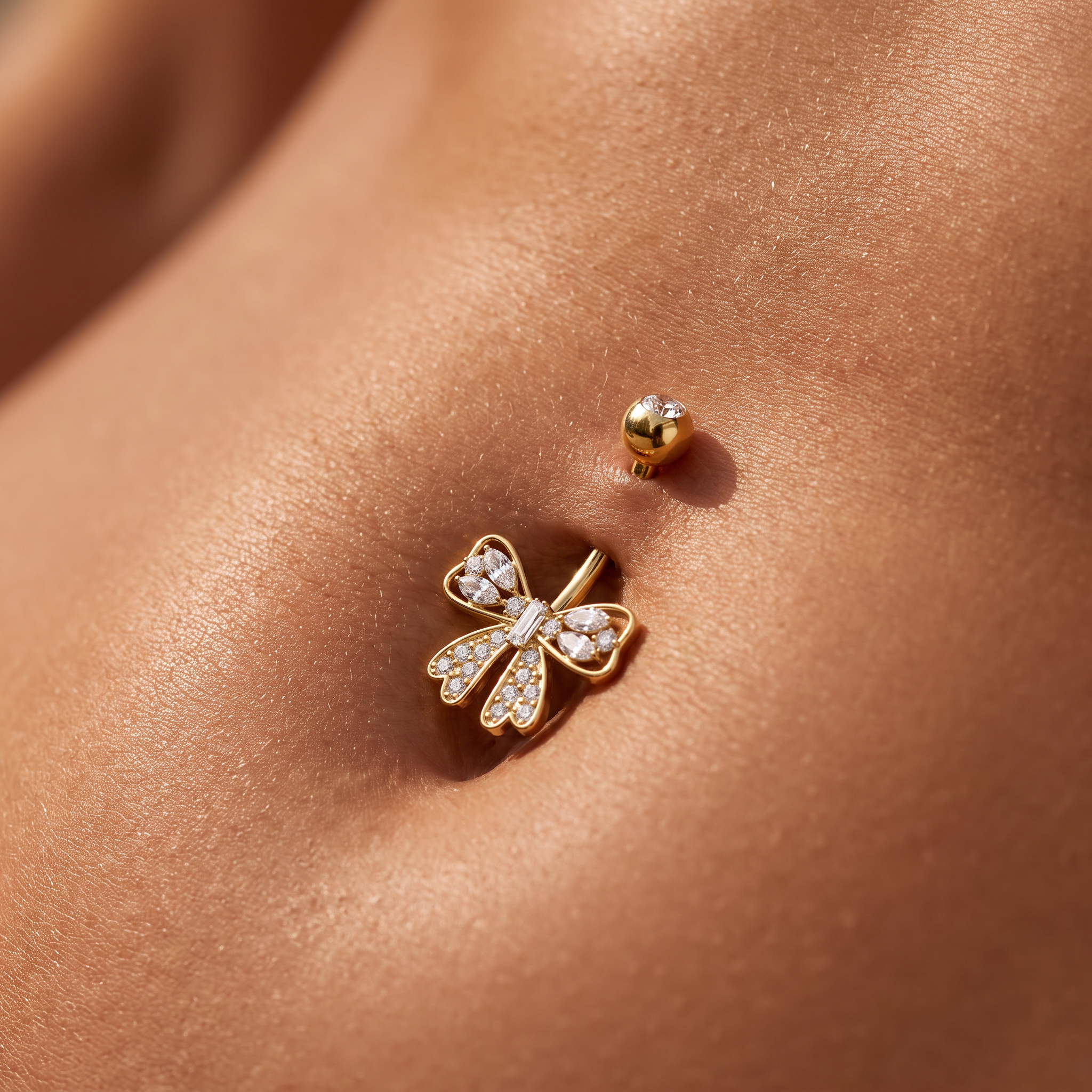 A close-up of a gold dragonfly stud piercing with clear pavé stones, shown worn on a person's navel with a matching gold ball stud above it.