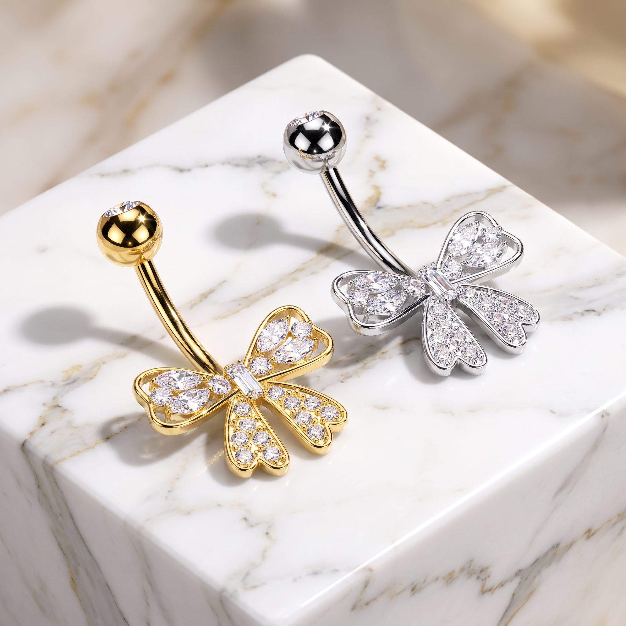 Two bow-shaped belly button rings displayed on a white marble cube. One ring is yellow gold with clear crystal pavé detailing, and the other is silver with matching clear crystal pavé detailing.