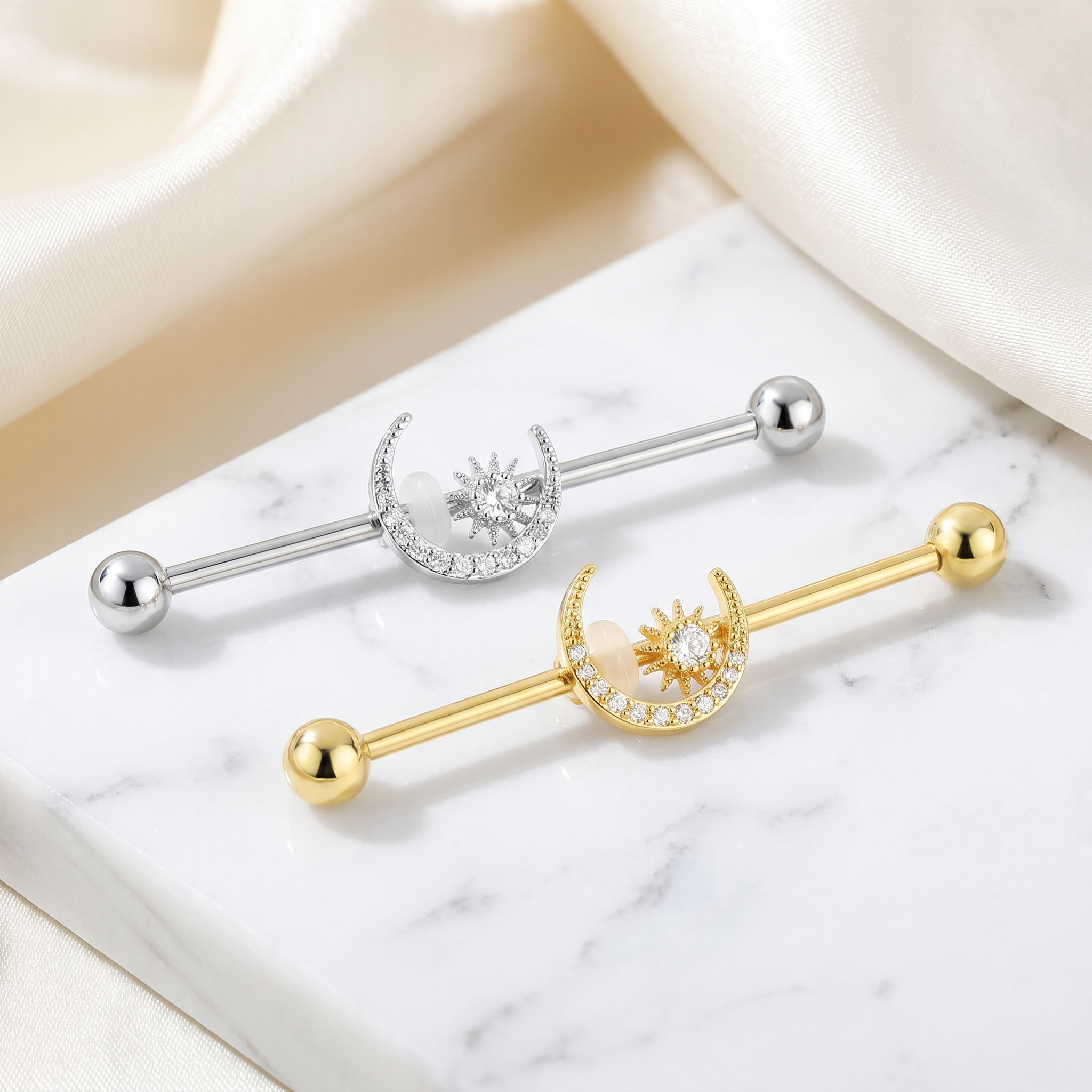 Two barbell nipple piercings with crescent moon and sun designs are displayed on a white marble slab with cream satin fabric in the background. One is silver-toned and the other is gold-toned, both featuring clear rhinestone detailing.