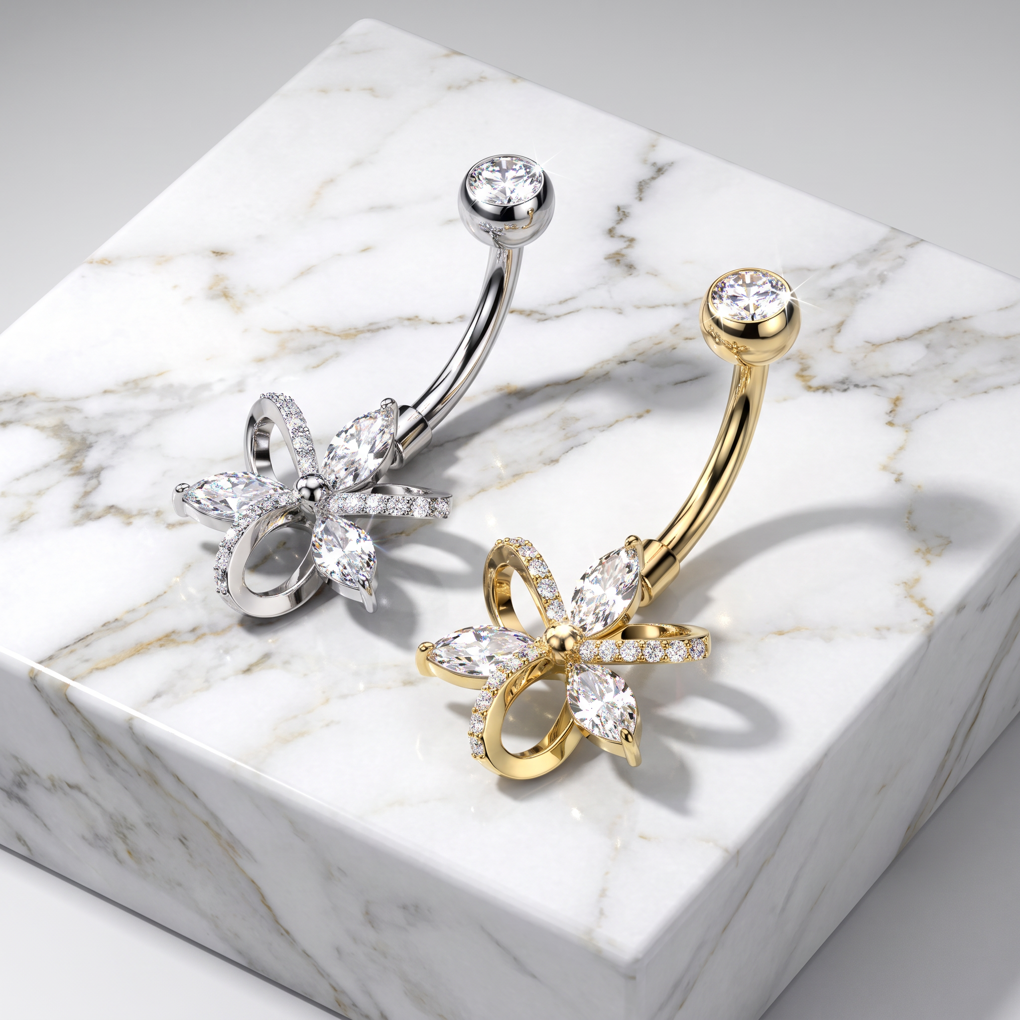 Two curved barbell belly button piercings with flower-shaped rhinestone charms, one in silver and one in gold, displayed on a white marble block.