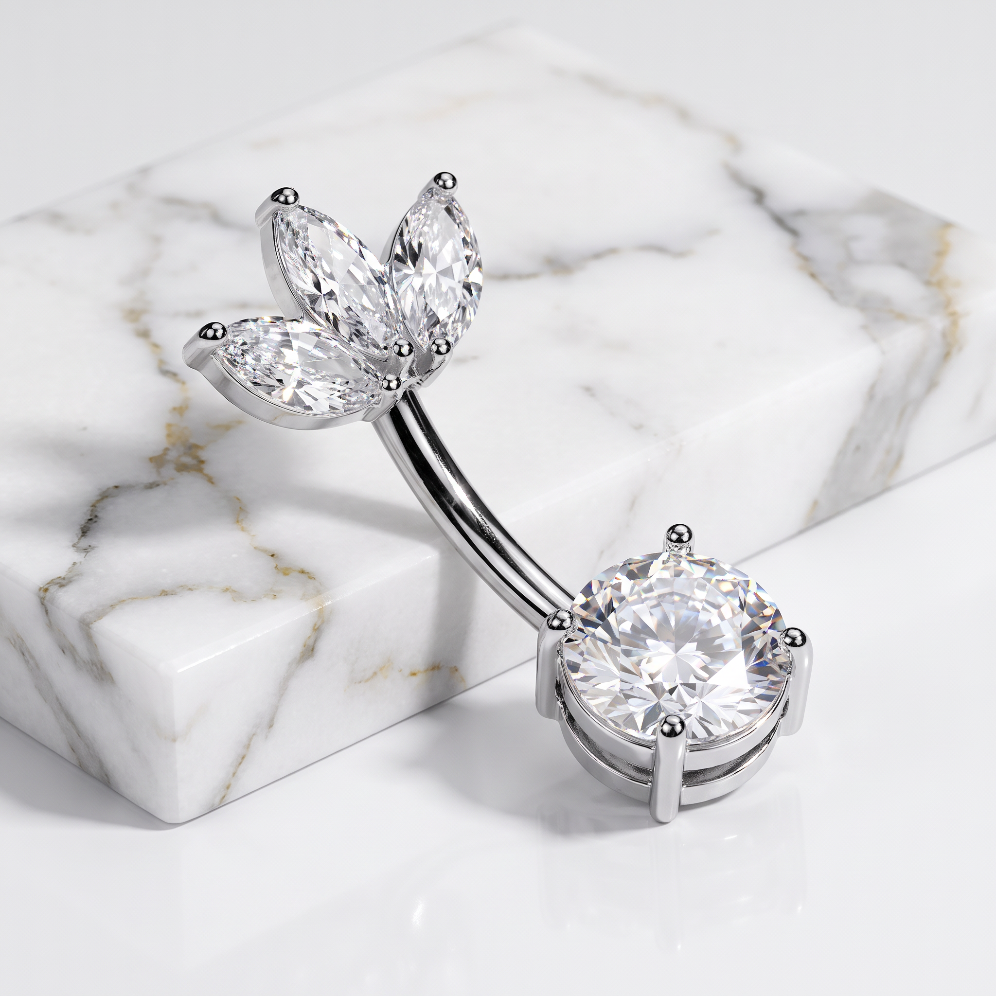 A silver-toned curved barbell belly button ring with marquise-cut clear gemstones on one end and a round clear gemstone on the other, displayed on a white marble block against a white background.