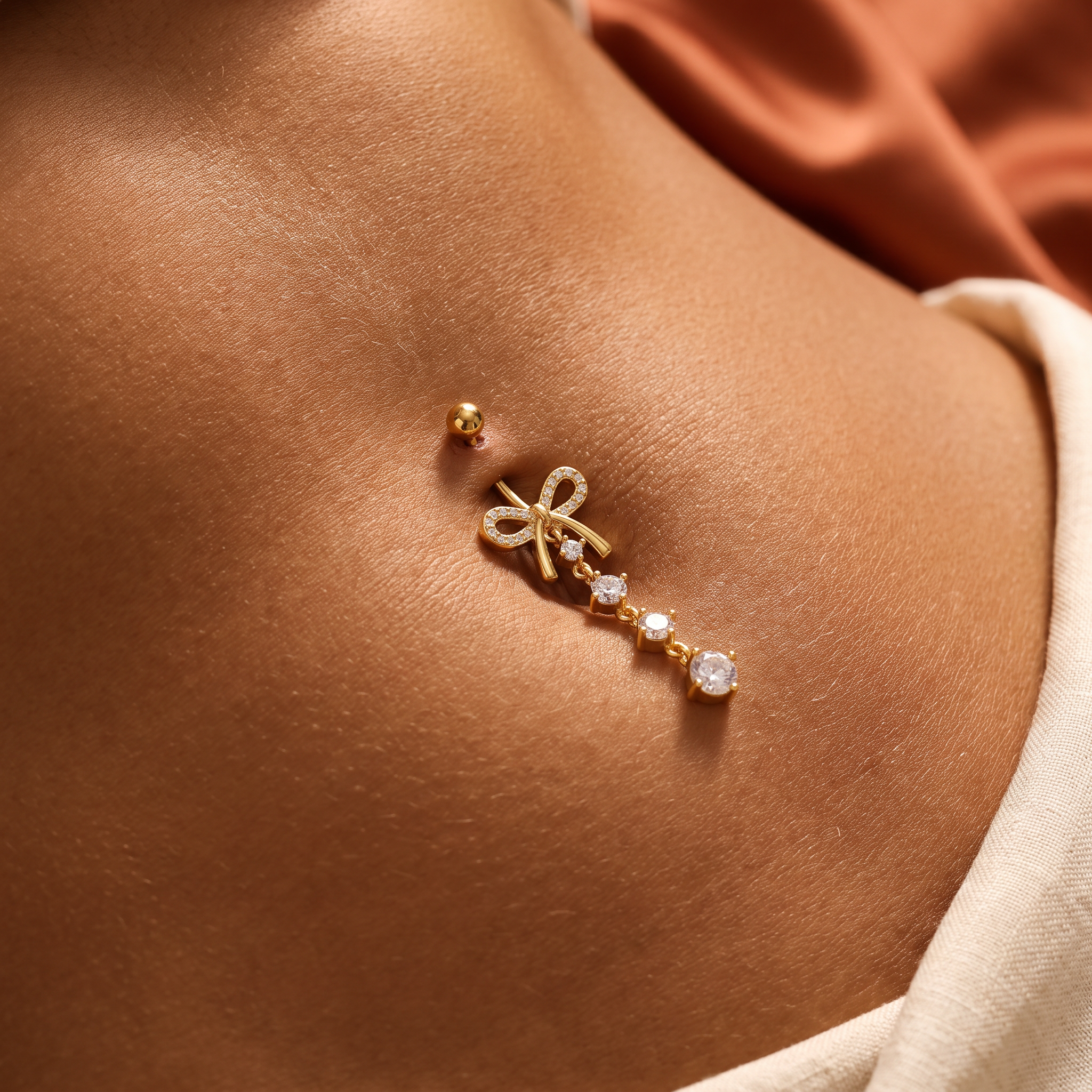 A close-up of a person's chest wearing a delicate gold belly button ring with a bow design and small clear gemstones, next to a matching gold stud earring.