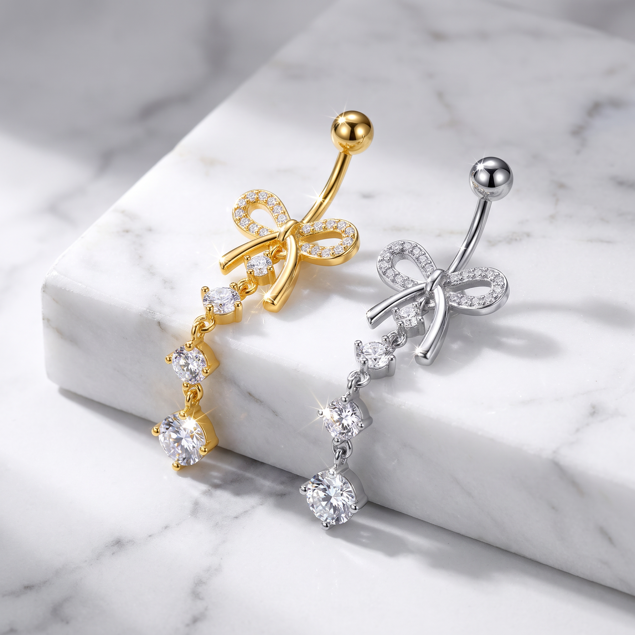 Two belly button rings with bow and dangling crystal designs, one in gold and one in silver, displayed on a white marble block.