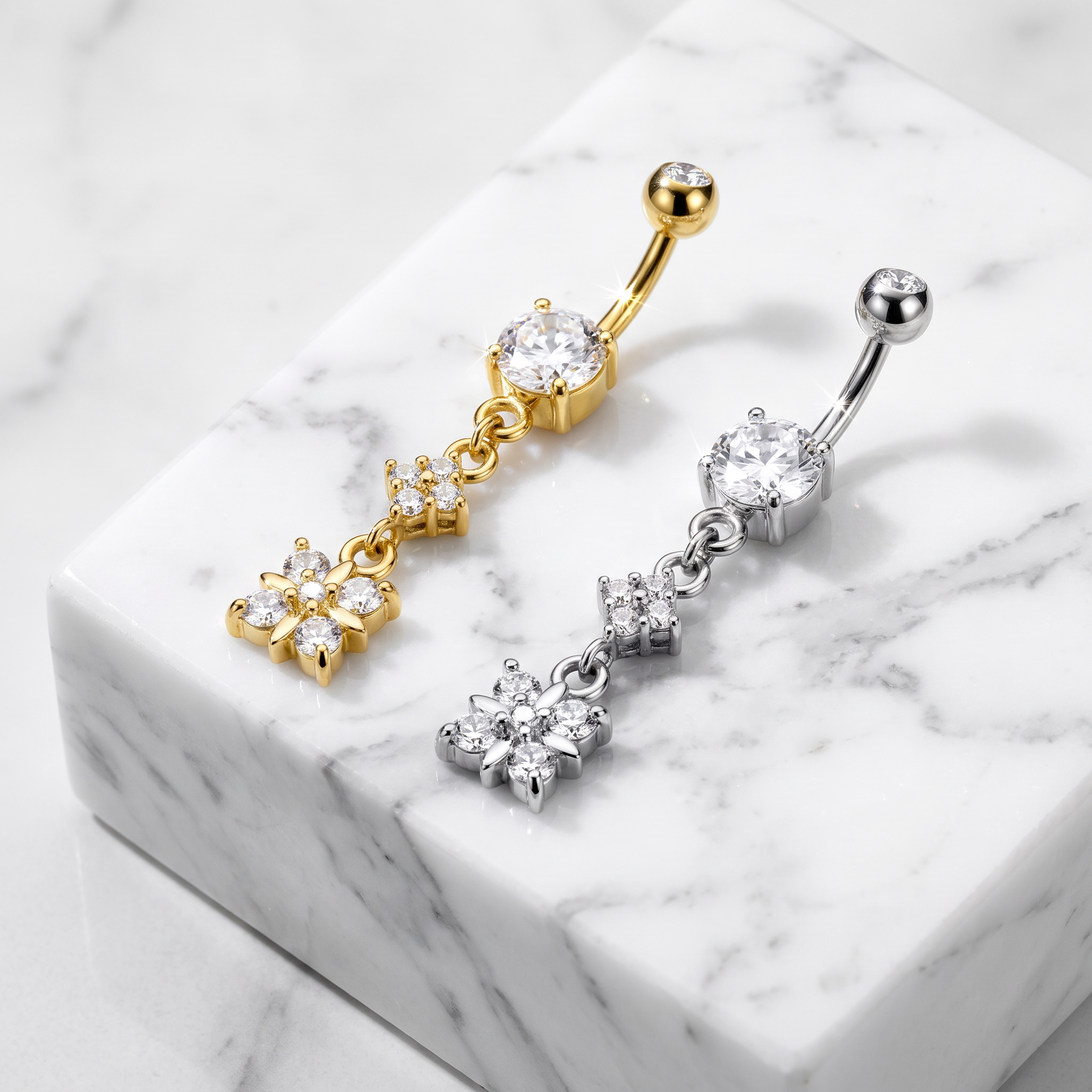 Two belly button rings with dangling floral and chain details, one in yellow gold and one in silver, displayed on a white marble block.