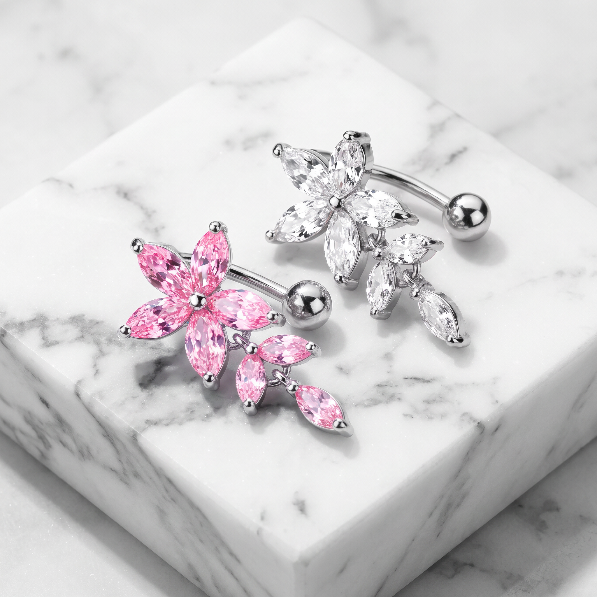 Two flower-shaped barbell belly button piercings displayed on a white marble block. One features pink marquise-cut crystals, and the other features clear crystals.