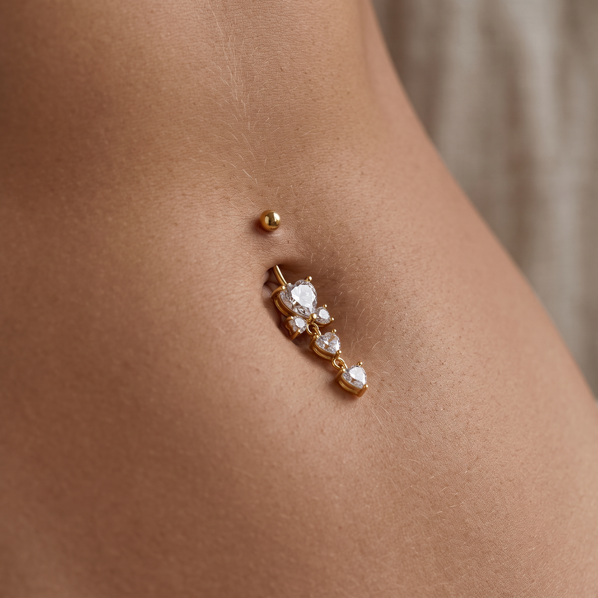A close-up of a gold navel piercing with a dangling cluster of clear cubic zirconia stones, shown on a model's lower abdomen.