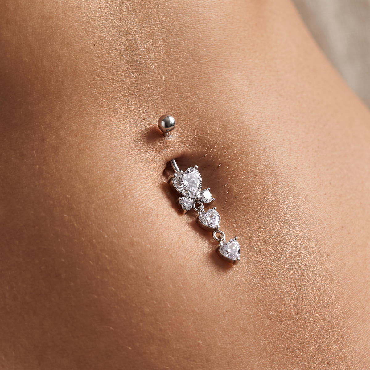 A close-up of a person's navel wearing a silver-toned belly button ring with a dangling floral cluster made of clear stones, alongside a simple matching stud above it.