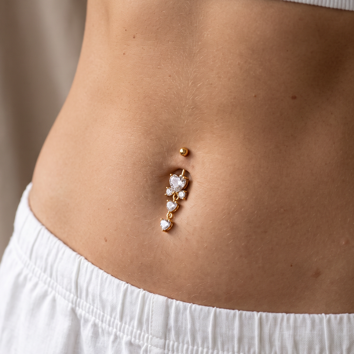 A close-up of a person's midsection wearing a gold belly button ring with clear rhinestones and small pearl-like accents, paired with a white ribbed crop top and white pants.