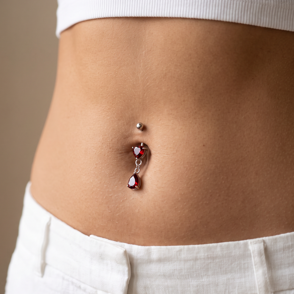 A close-up of a person's lower torso wearing a silver belly button ring with dangling red heart and teardrop gemstones, paired with a white ribbed crop top and white pants.