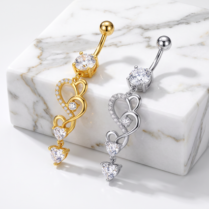 Two heart-themed belly button rings, one in gold and one in silver, both featuring a large round clear stone and three cascading heart designs with small clear stones, displayed on a white marble block.