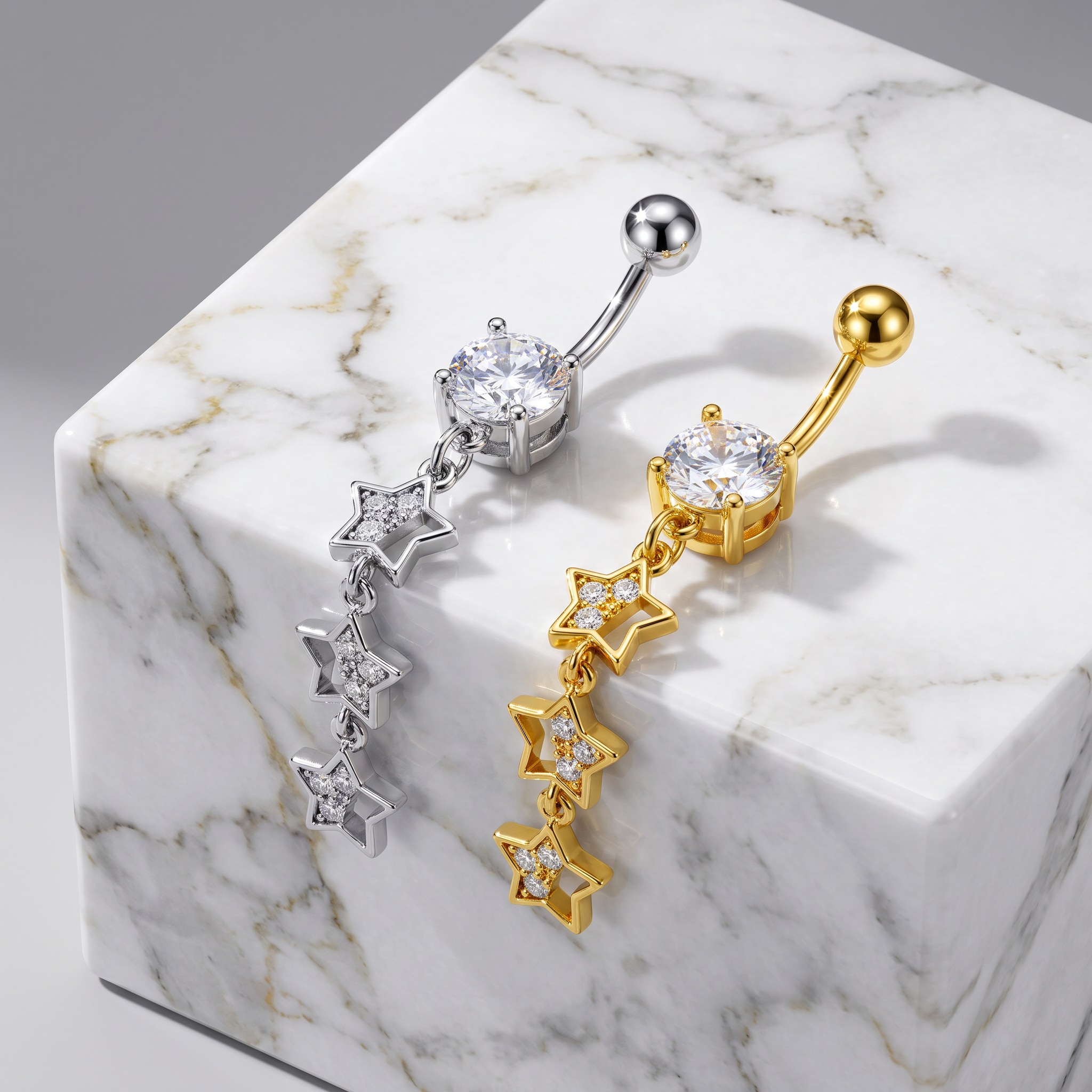 Two belly button rings with star-shaped dangling charms are displayed on a white marble block. One ring is silver-toned with clear gemstones, and the other is gold-toned with clear gemstones.