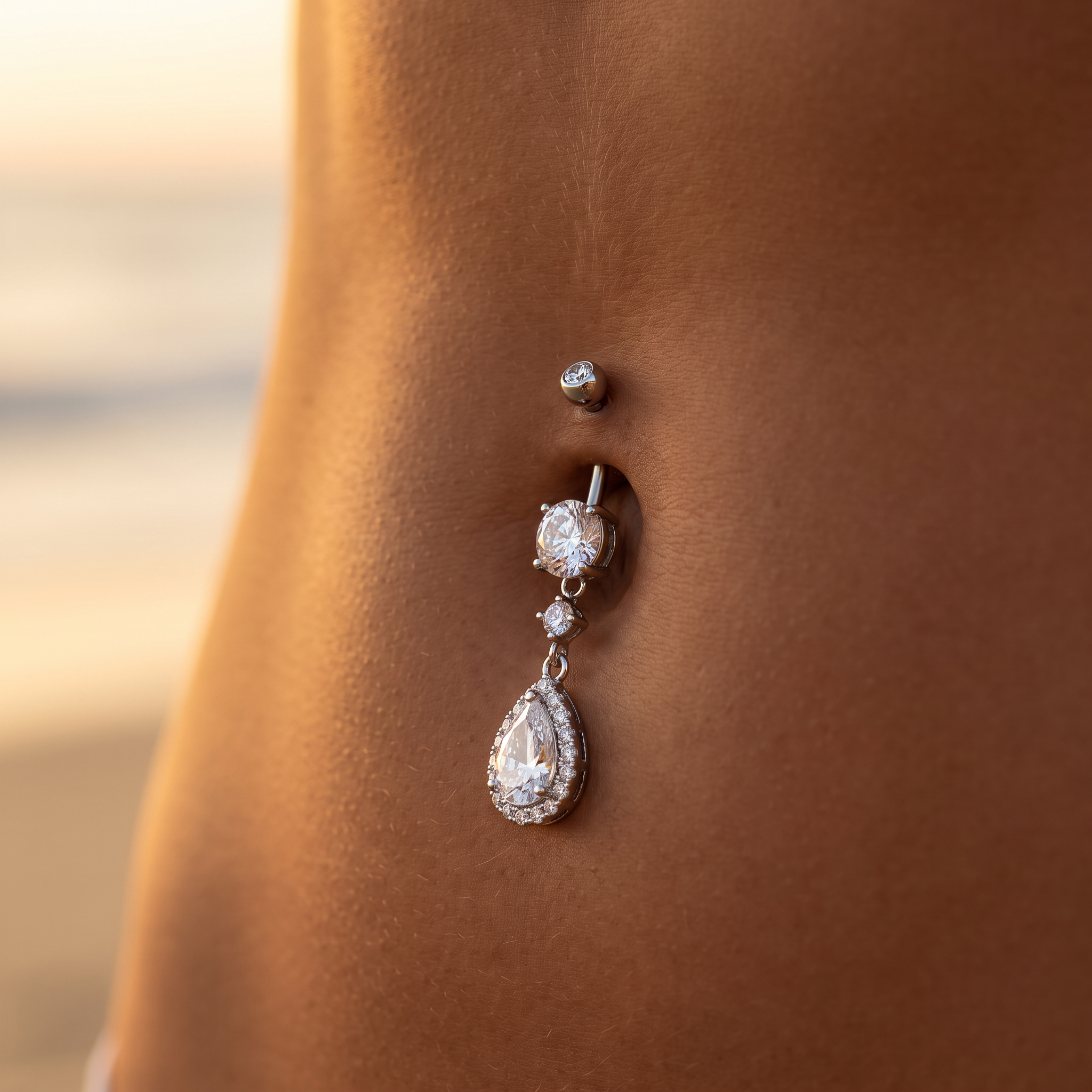 A silver-toned belly button ring with a teardrop-shaped clear gemstone and smaller clear stones, shown worn on a person's abdomen against a softly blurred beach background.