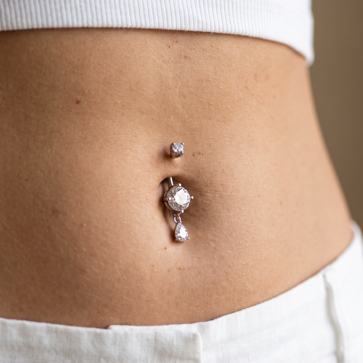 A close-up of a person's lower torso showing a belly button piercing with a silver-toned vertical barbell stud featuring a clear teardrop-shaped gemstone, worn with a white ribbed crop top and white pants.