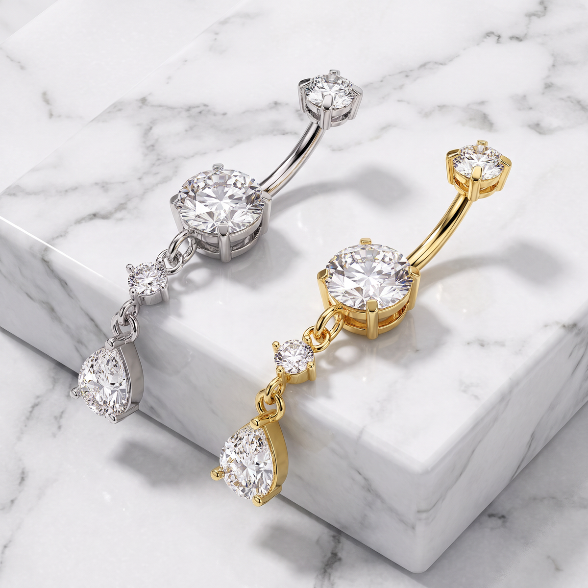 Two curved barbell belly button piercings with large round and teardrop-cut clear gemstones, shown on a white marble block. One is silver-toned and the other is yellow gold-toned.