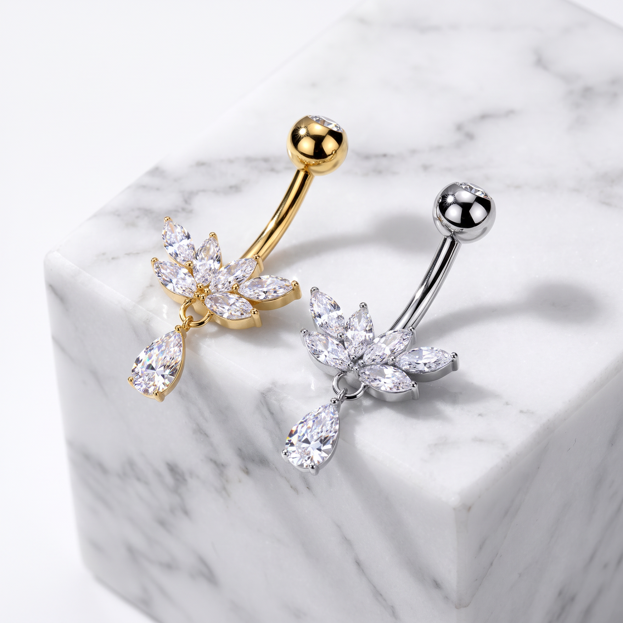 Two flower-shaped belly button rings with clear crystal petals, one in gold-tone metal and one in silver-tone metal, displayed on a white marble cube.