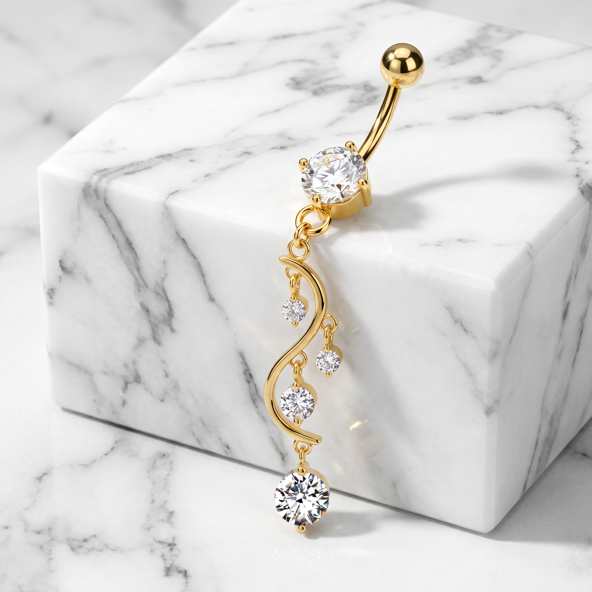A gold belly button ring with a dangling wavy chain and clear cubic zirconia stones, displayed on a white marble block over a marble surface.
