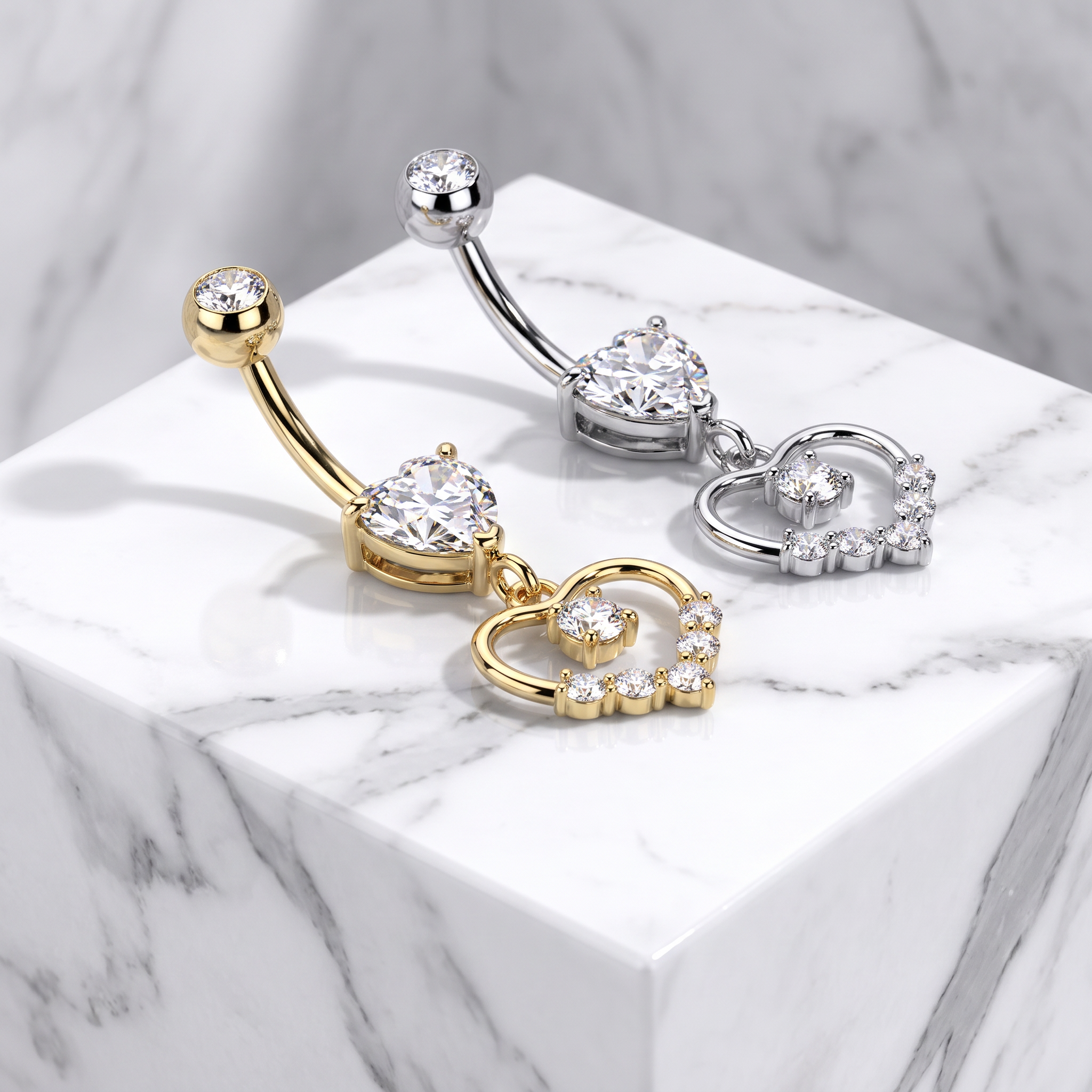 Two curved barbell belly button piercings with heart-shaped crystal clusters and dangling heart charms, shown in gold and silver on a white marble pedestal.