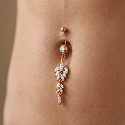 A gold belly button ring with a dangling leaf cluster design set with clear crystals, shown worn on a person's abdomen.