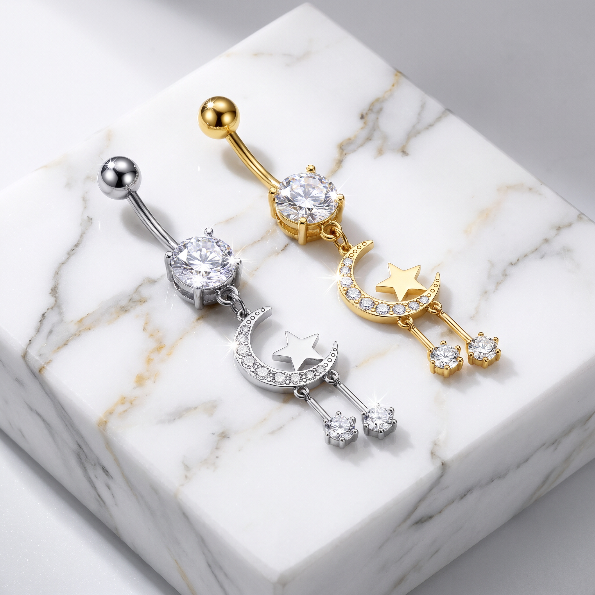 Two celestial belly button rings displayed on a white marble block. One ring is silver-toned with a moon, star, and dangling rhinestone charms, and the other is gold-toned with matching celestial details.