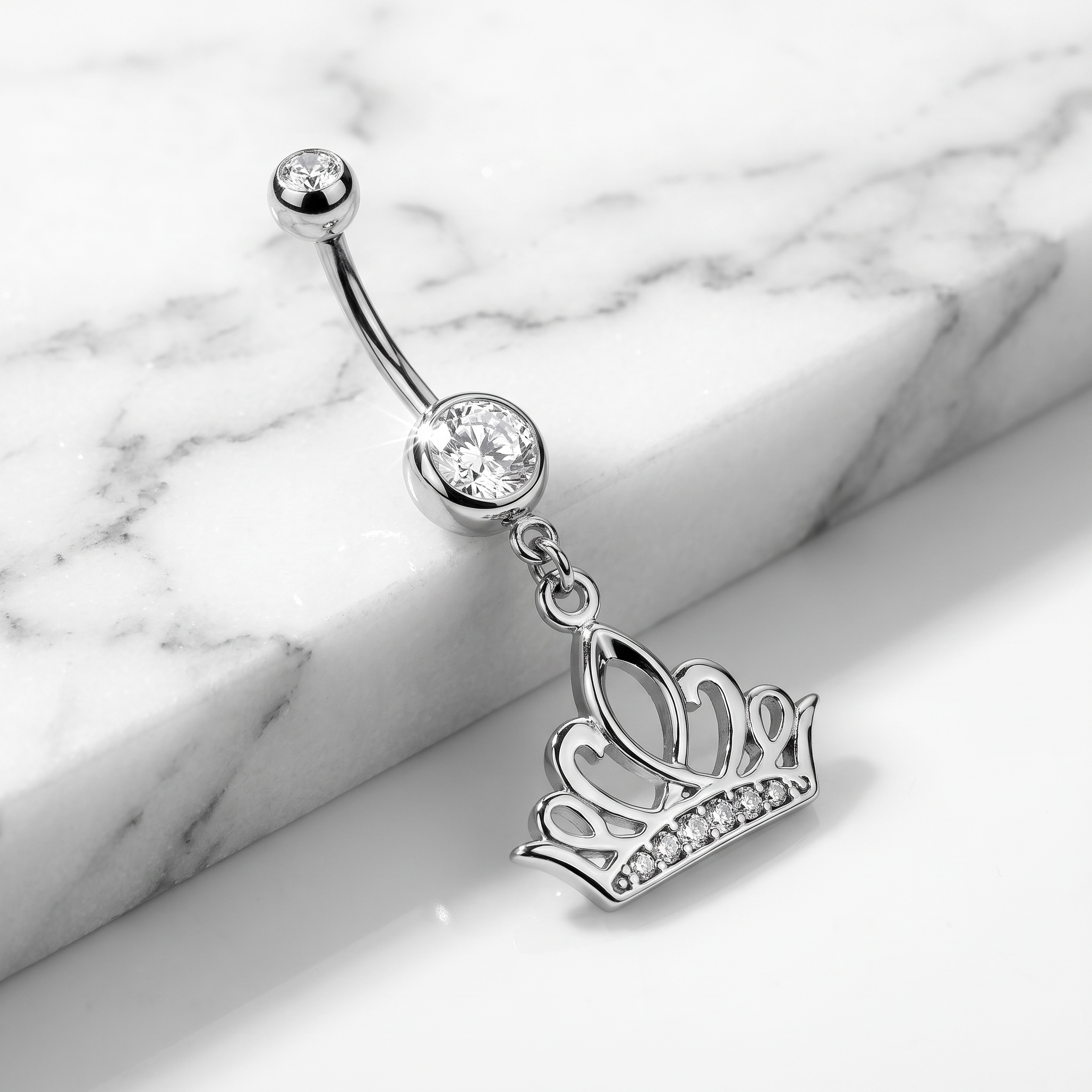 A silver-toned belly button ring with a curved barbell, a round clear gemstone, and a dangling crown charm adorned with small clear stones, displayed on a white marble surface.