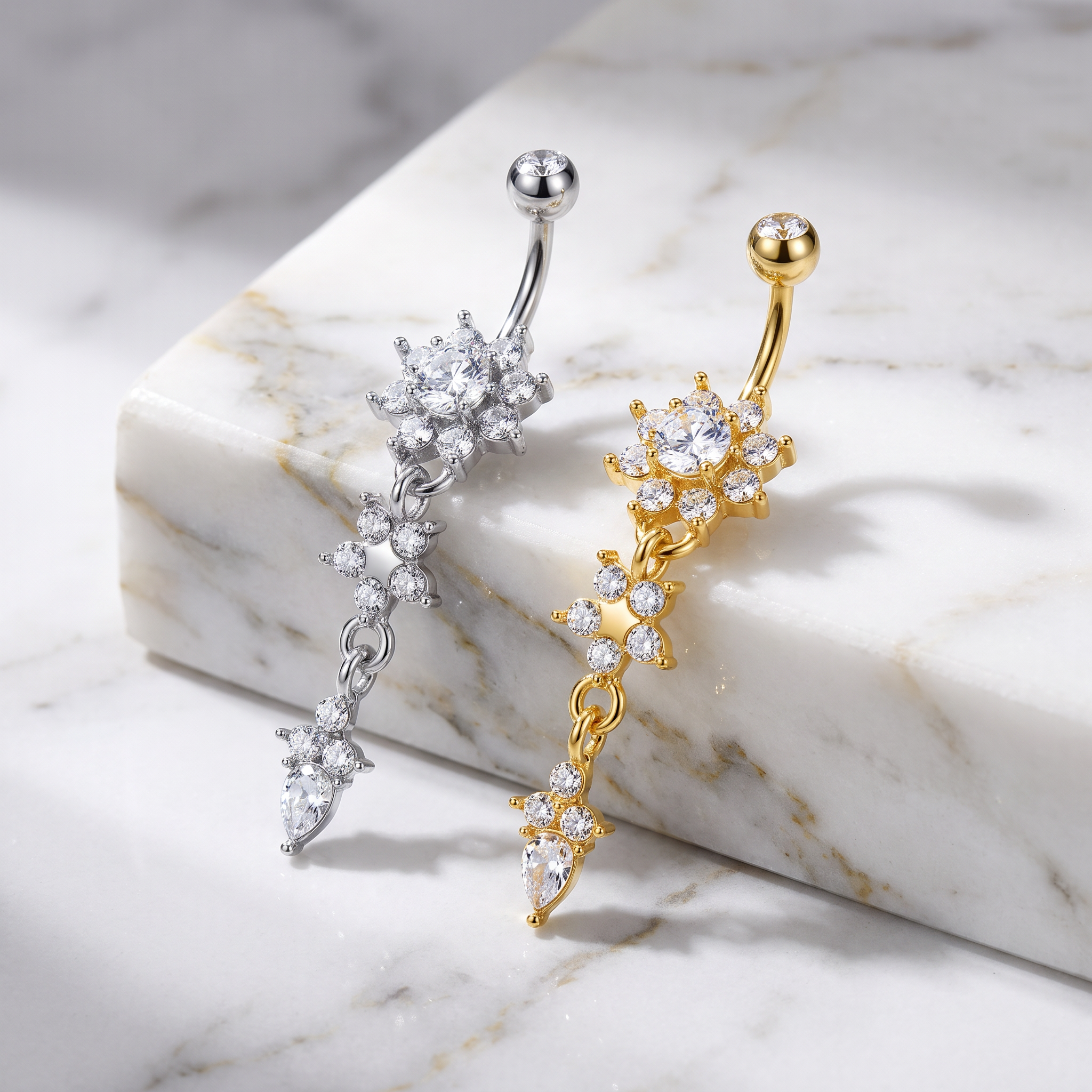Two belly button rings with floral and chain-like crystal clusters, one in silver-tone metal and one in gold-tone metal, displayed on a white marble block over a light marble surface.