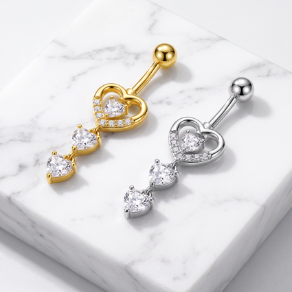 Two heart-shaped dangling belly button rings, one in yellow gold and one in silver, both featuring clear heart-shaped gemstones, displayed on a white marble block.