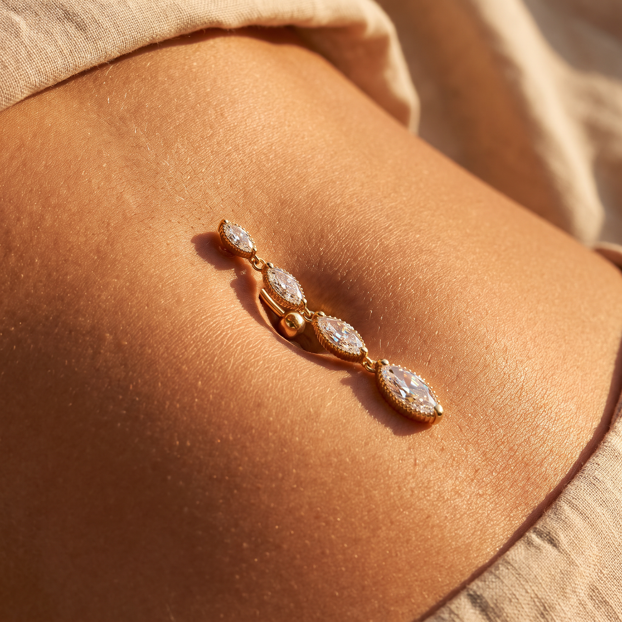 A gold belly button ring with three marquise-cut clear stones, shown worn on a person's navel against a warm beige background.