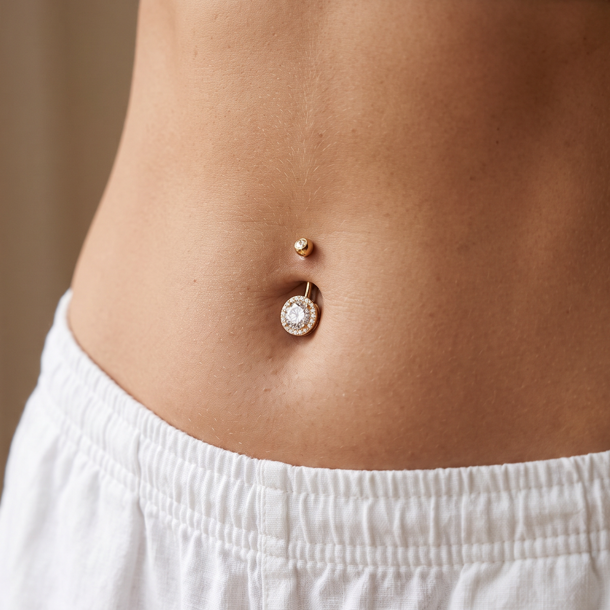 A close-up of a person's midsection wearing a gold belly button ring with a small round stud and a dangling charm with a clear rhinestone, styled with white clothing.