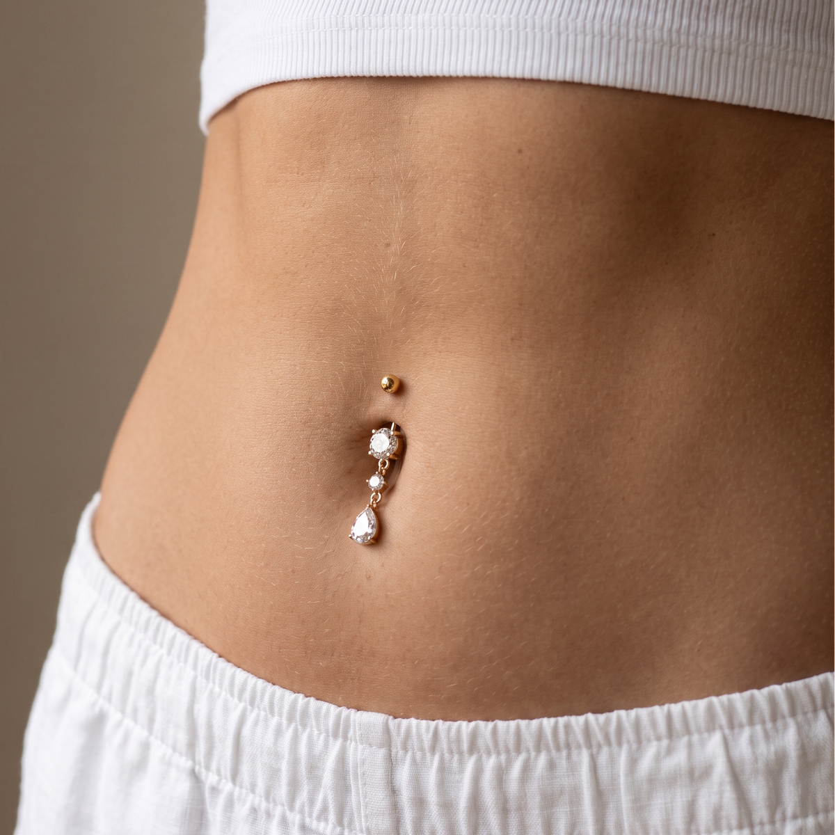 A close-up of a person's lower torso wearing a white ribbed crop top and white pants, featuring a belly button ring with a gold ball, clear rhinestones, and a teardrop pearl.