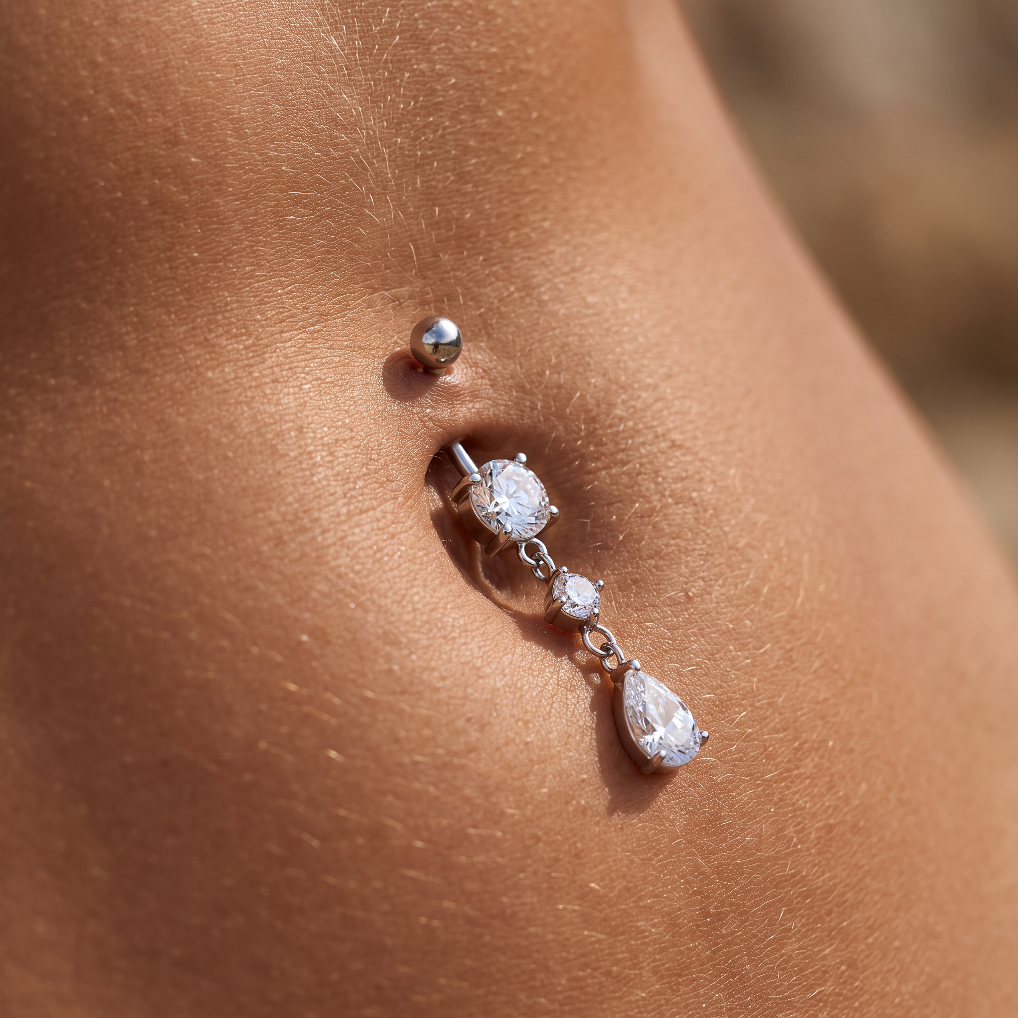 A close-up of a silver-toned navel piercing with a curved barbell and clear teardrop and round stones, shown on human skin.