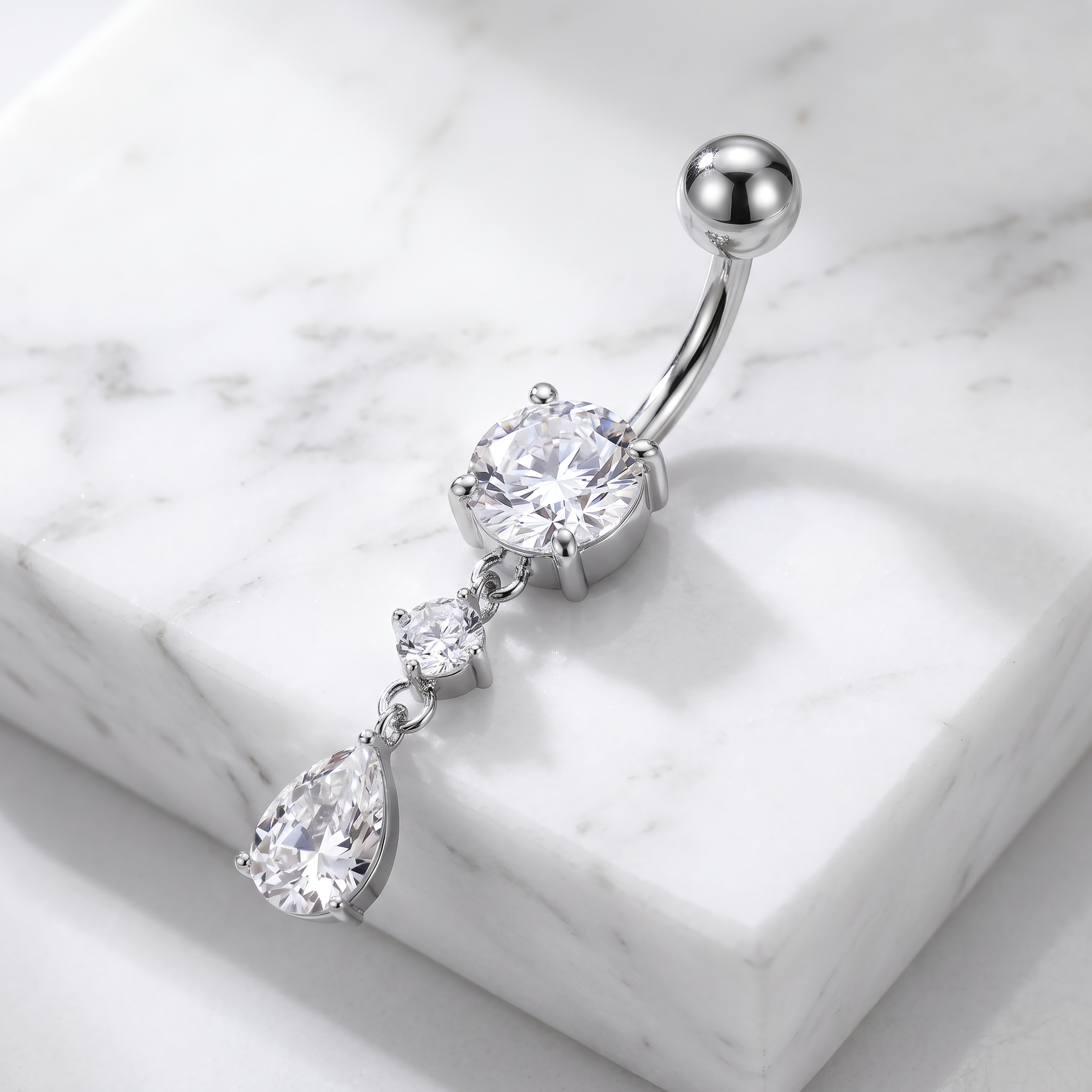 A silver-toned curved barbell belly button ring with a large round clear gemstone and two dangling teardrop-shaped clear gemstones, displayed on a white marble block.