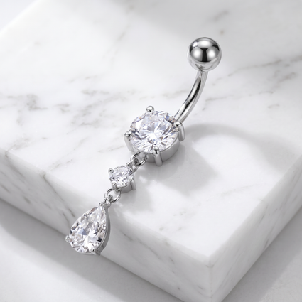 A silver-toned curved barbell belly button ring with a large round clear gemstone and two dangling teardrop-shaped clear gemstones, displayed on a white marble block.