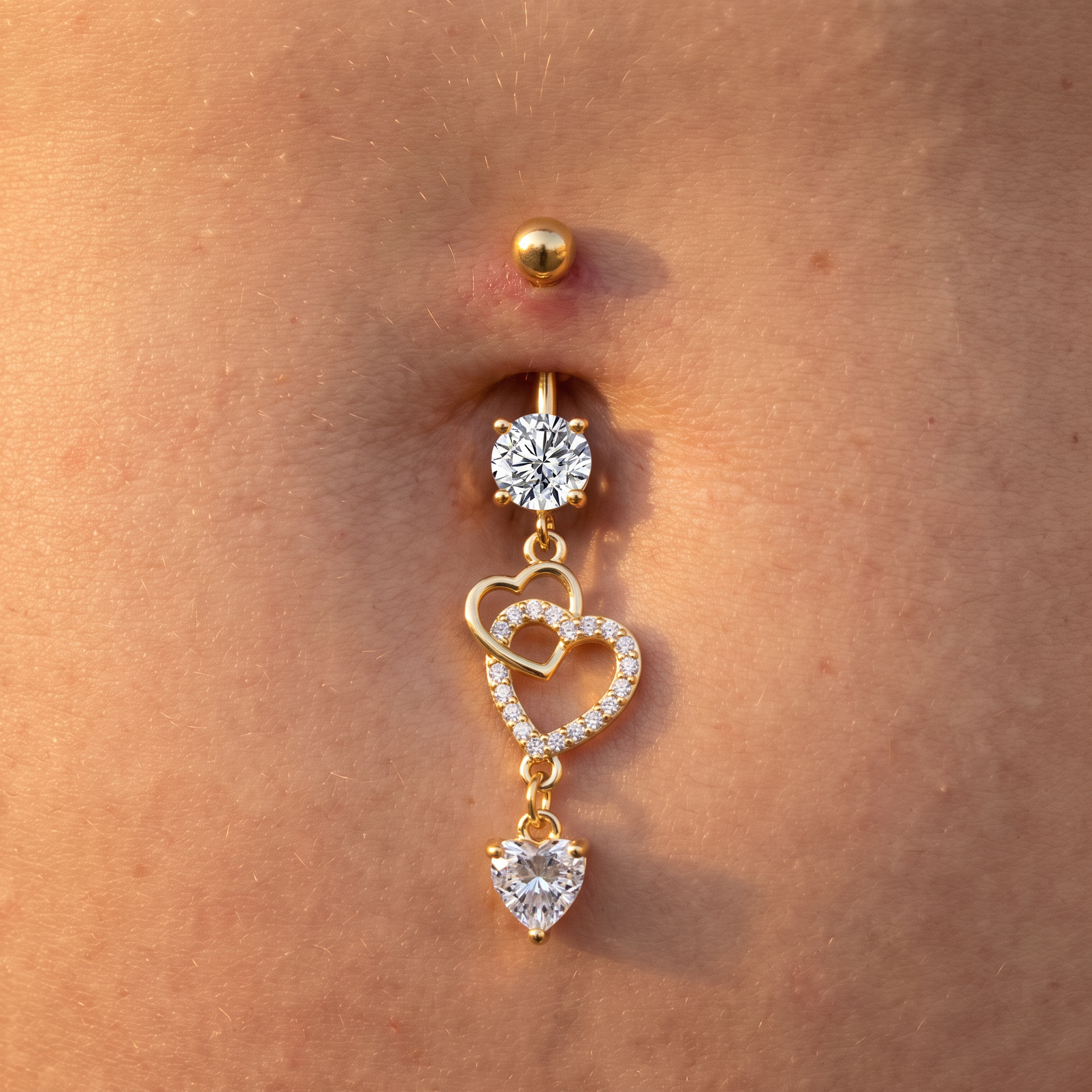 Gold belly button ring with a round clear gemstone, interlocking heart charms outlined with small clear stones, and a dangling heart charm, shown worn on a person's abdomen.