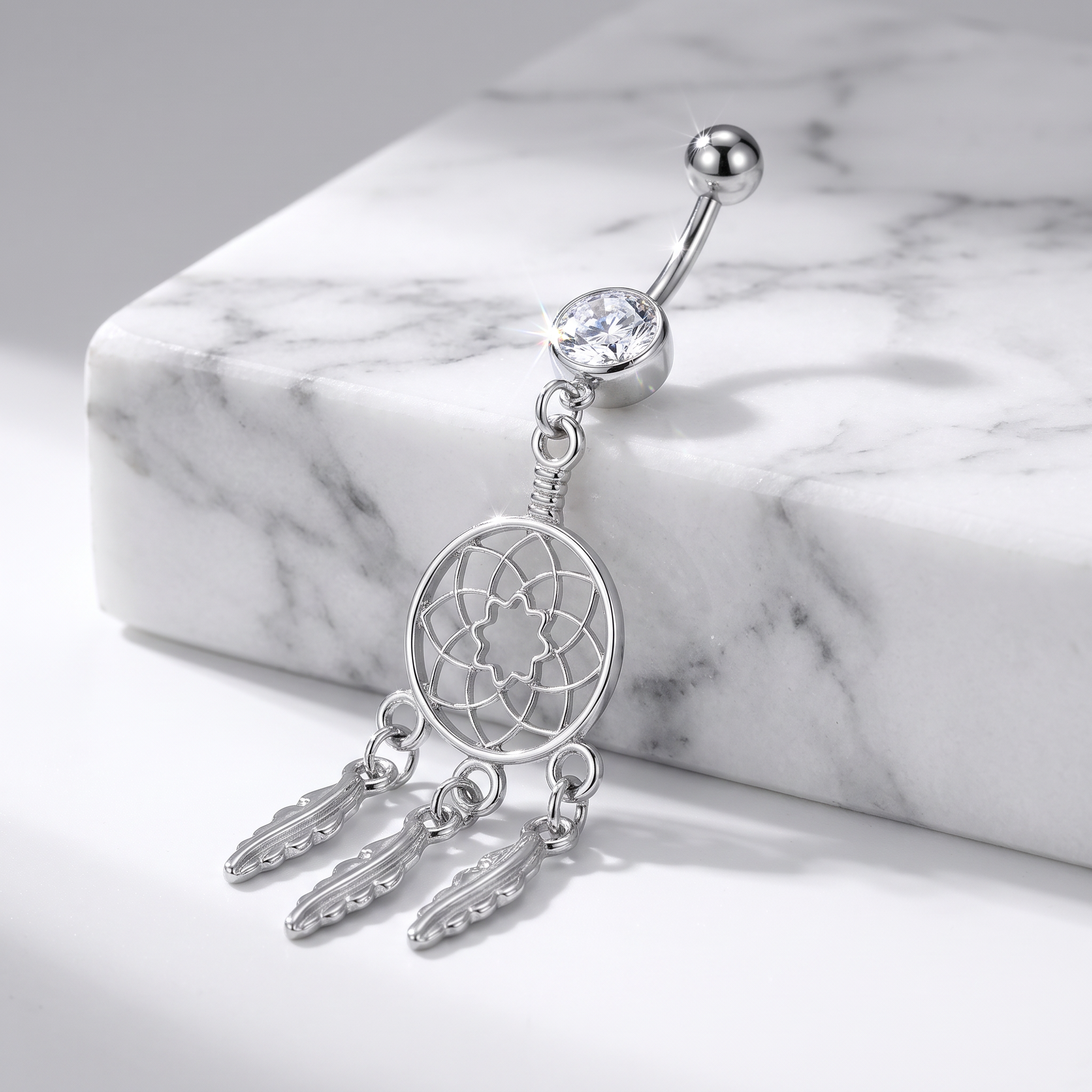 A silver dreamcatcher belly button ring with dangling feather charms and a clear rhinestone, displayed on a white marble block.