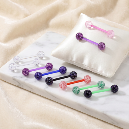 Several colorful barbell-style body jewelry pieces with glittery round ends are arranged on a white marble slab and a white satin pillow against a soft beige fabric background.