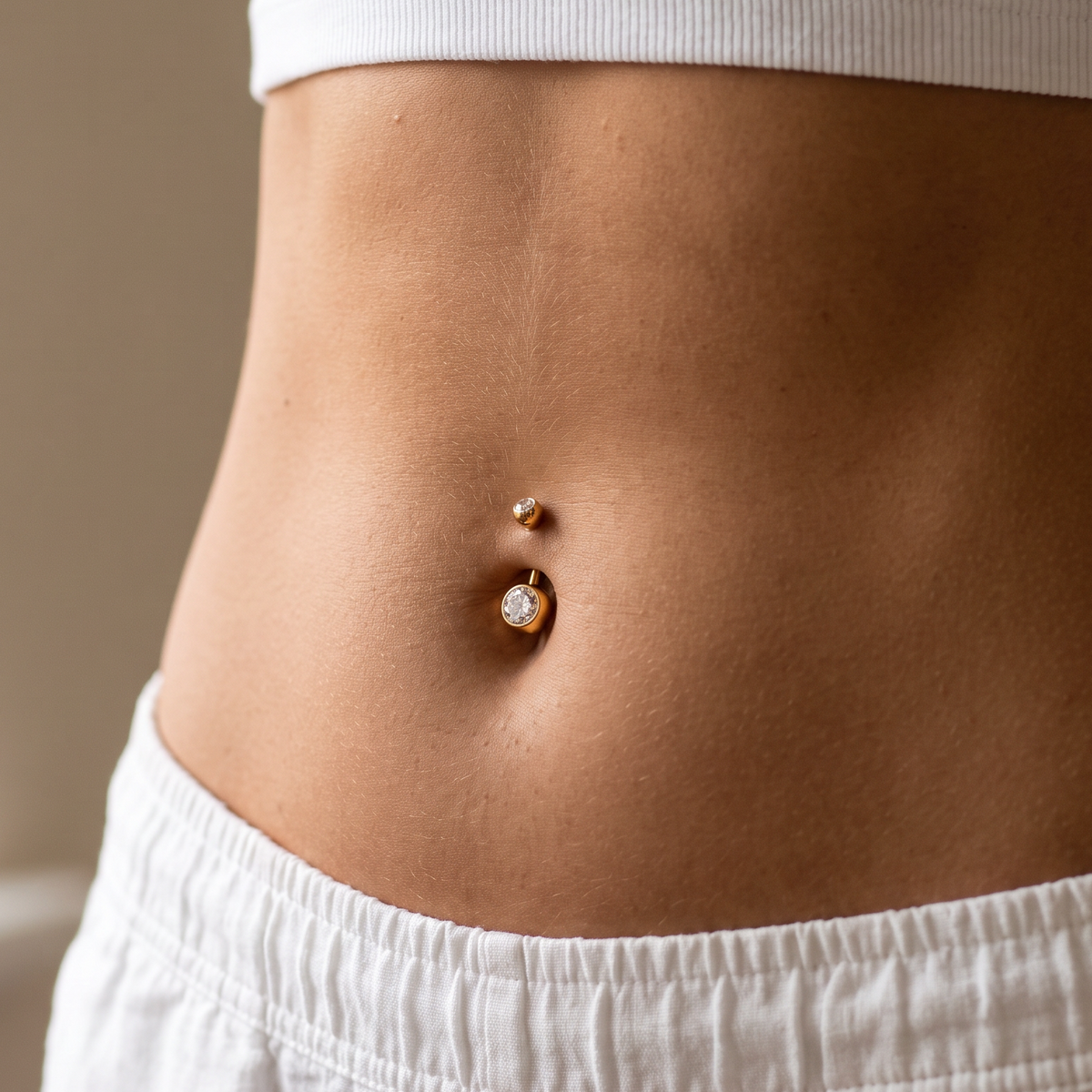 A close-up of a person's midsection wearing a white ribbed crop top and white shorts, featuring a gold belly button ring with a small clear gemstone.