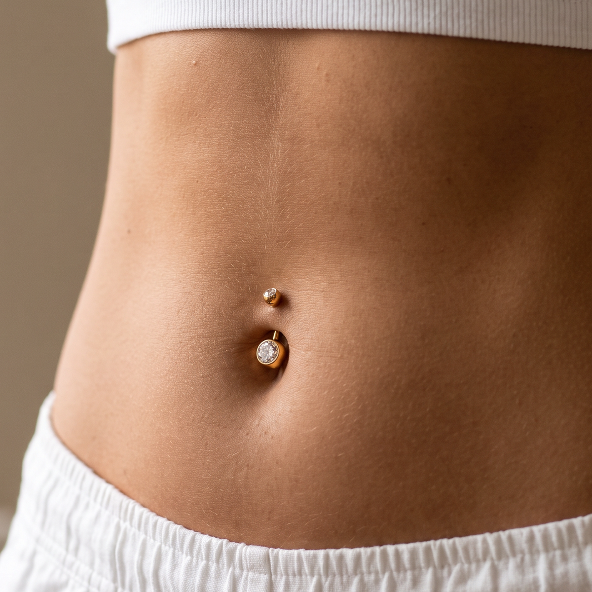 A close-up of a person's midsection wearing a white ribbed crop top and white shorts, featuring a gold belly button ring with a small clear gemstone.