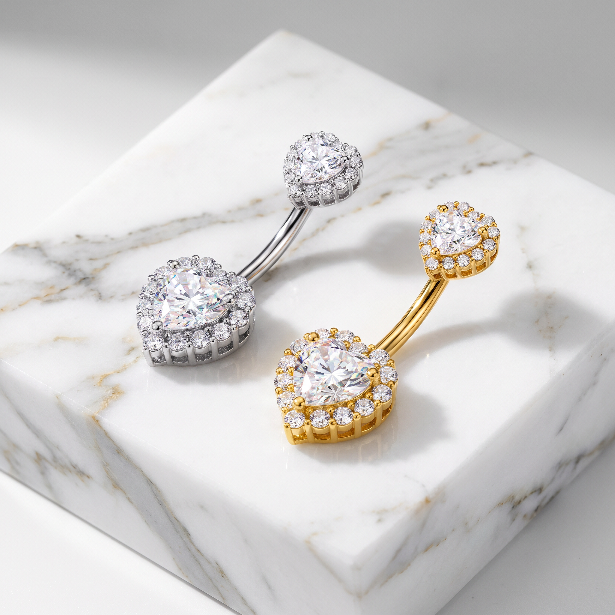 Two curved barbell belly button rings with halo-set round clear stones, one in silver-toned metal and one in yellow-gold metal, displayed on a white marble block.