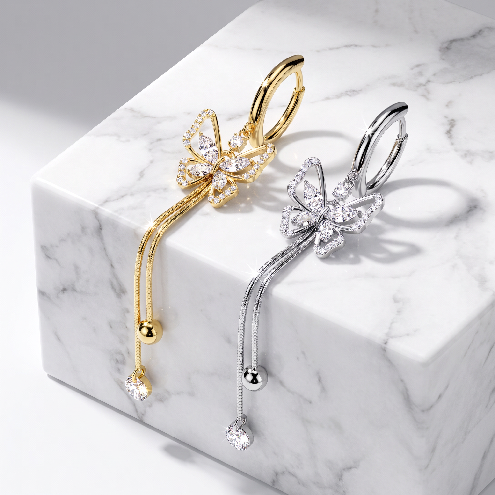 Two butterfly-shaped hoop earrings displayed on a white marble block, one in yellow gold and one in silver, both adorned with clear crystal pavé detailing and dangling crystal drops.