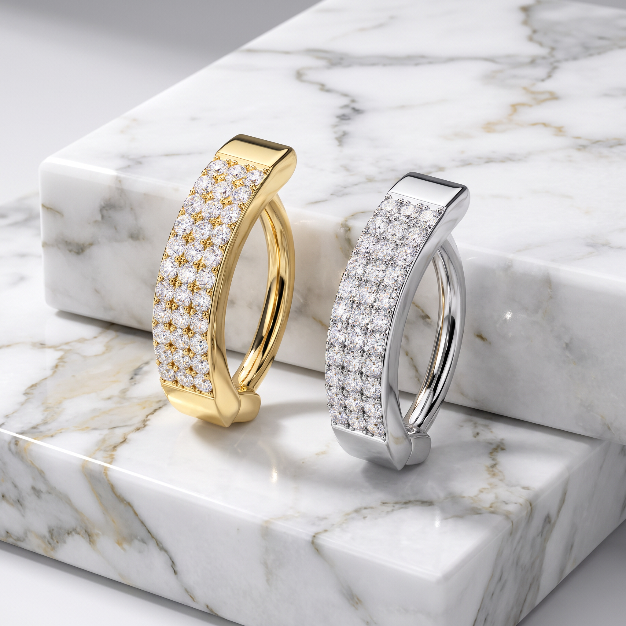 Two curved hoop-style earrings displayed on a white marble slab. One earring is yellow gold with pavé-set clear stones, and the other is silver-toned with pavé-set clear stones.