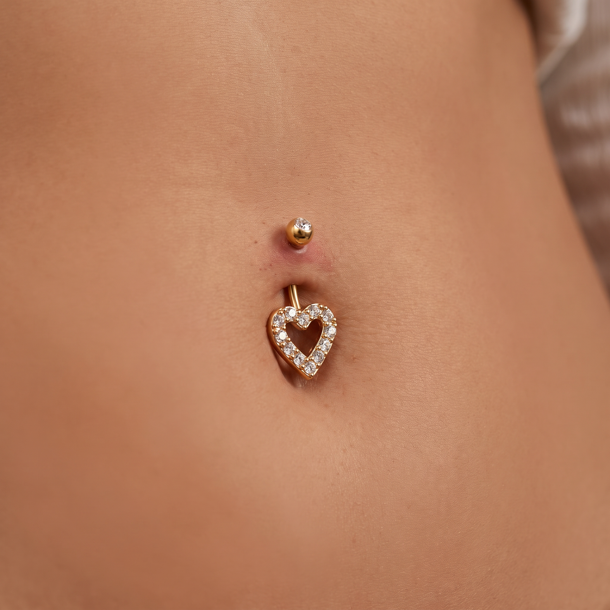 A close-up of a person's lower torso showing a gold belly button ring with a heart-shaped crystal border and a small gold stud above it.