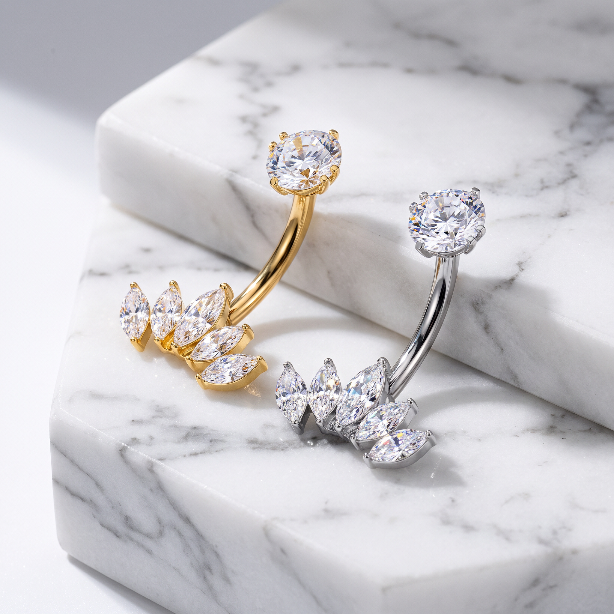 Two curved barbell-style body jewelry pieces with clear gemstones, one in yellow gold and one in silver, displayed on a white marble surface.
