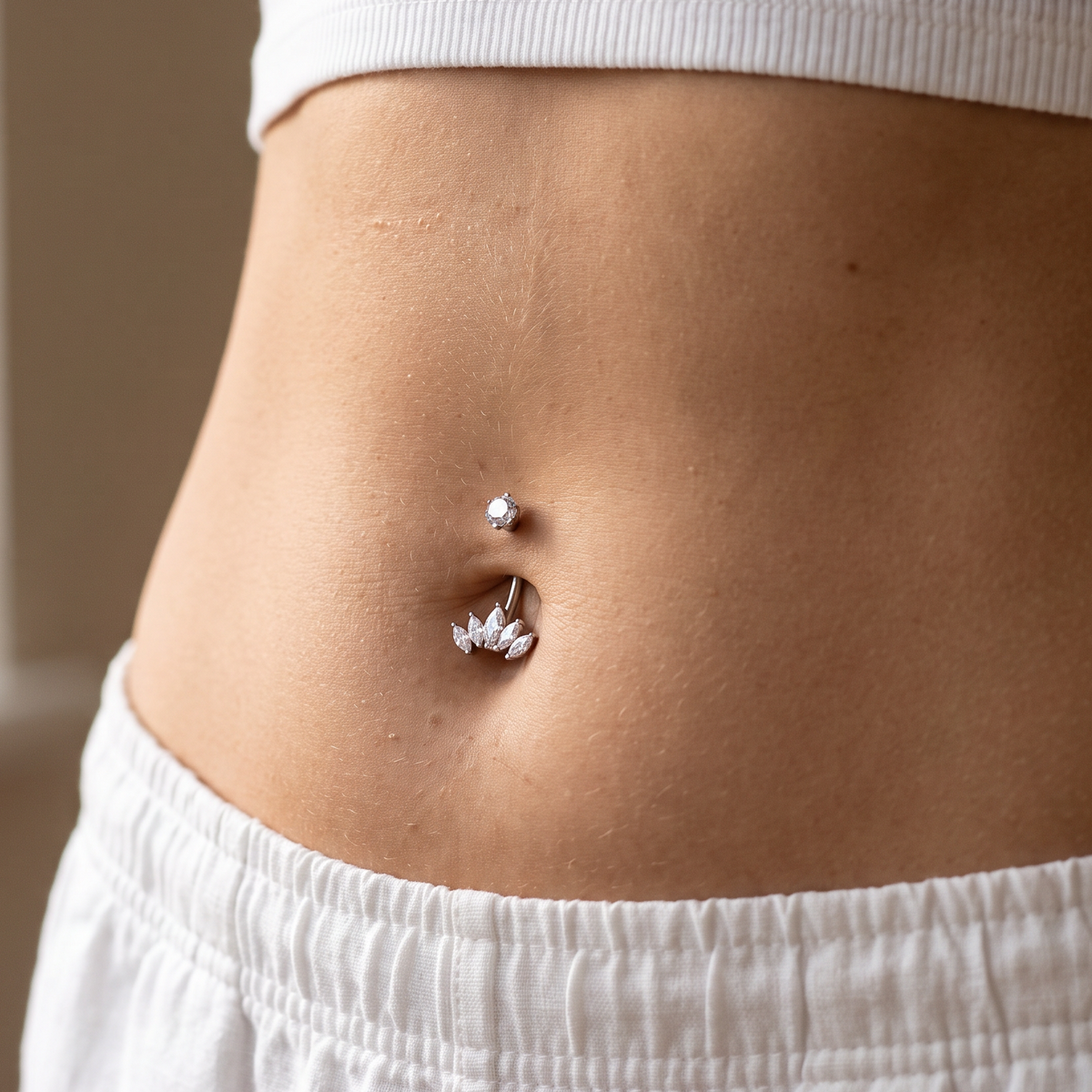 Close-up of a person's navel area wearing two belly button piercings: a small silver stud above and a silver hoop with a small flower-shaped charm below, styled with a white ribbed crop top and white pants.