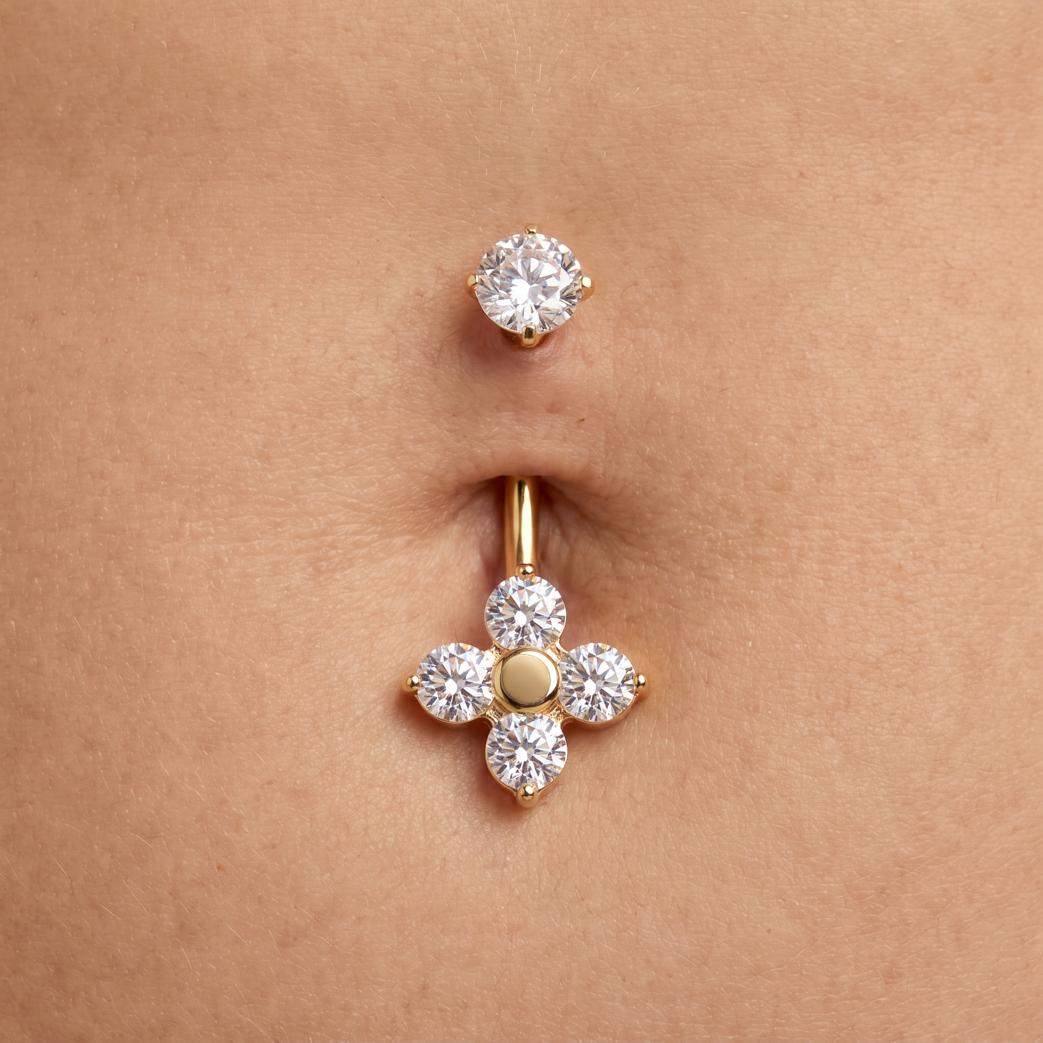 A close-up of a belly button with two gold piercings: a small stud above and a larger belly button ring below featuring a four-petal flower design with clear gemstones and a central gold dot, set against natural skin.